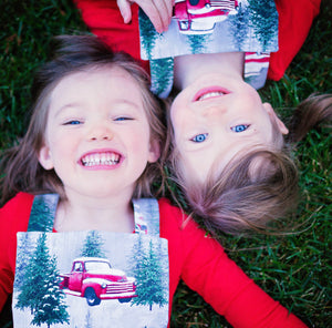 Sisters laying head to head in grass wearing Christmas pinafores: Baby holiday dresses featuring vintage trucks in the snow