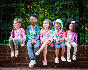 Tie dye baby clothes and kids tie dye clothing designs worn by five smiling kids on brick wall.  Sibling coordinating outfits by Worthy Threads clothing brand.