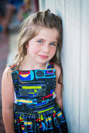 Girl modeling toddler pinafore dress in Boombox Print.  Unique kids clothing by Worthy Threads clothing brand.