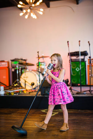 Girl singing into microphone wearing pinafore dinosaur dress.  Unique kids clothing available in matching sister outfits newborn and toddler!