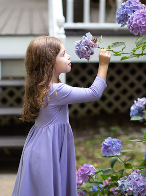 Toddler Girl modeling purple twirly dress while smelling purple flowers