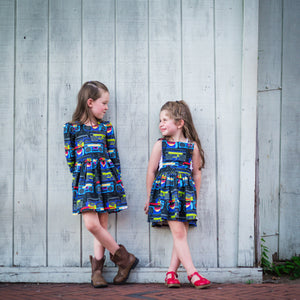 Two girls looking at each other wearing boombox print dresses.  Toddler & baby pinafore dress, retro baby clothes featured.  Matching sister outfits newborn and toddler available!