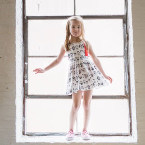 Baby pinafore dress in Beatnik print modeled by girl standing in windowsill.  Unique toddler clothes by Worthy Threads clothing brand with sibling coordinating outfits available!